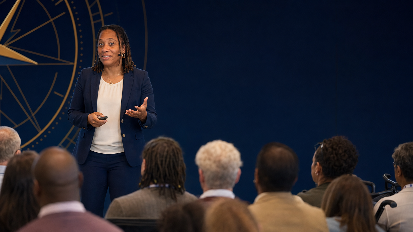Nichole Braxton speaks on stage before a diverse seated audience of educators, with a large gold compass rose on the navy wall behind her.