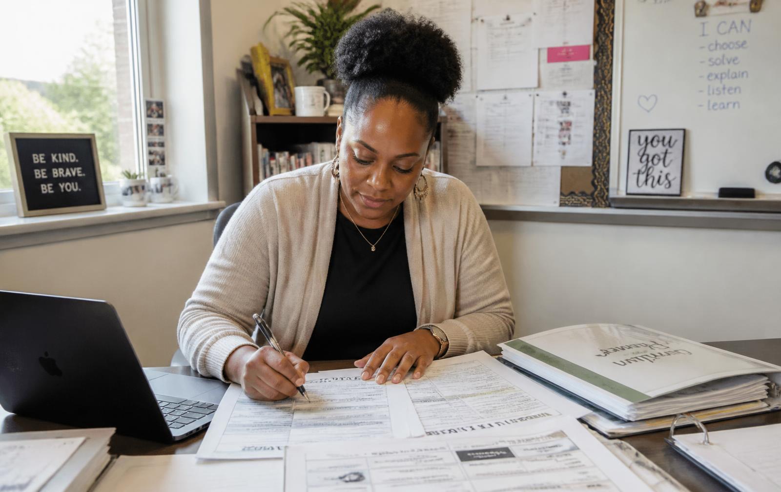 A teacher works through printed planning templates and a laptop at her classroom desk.